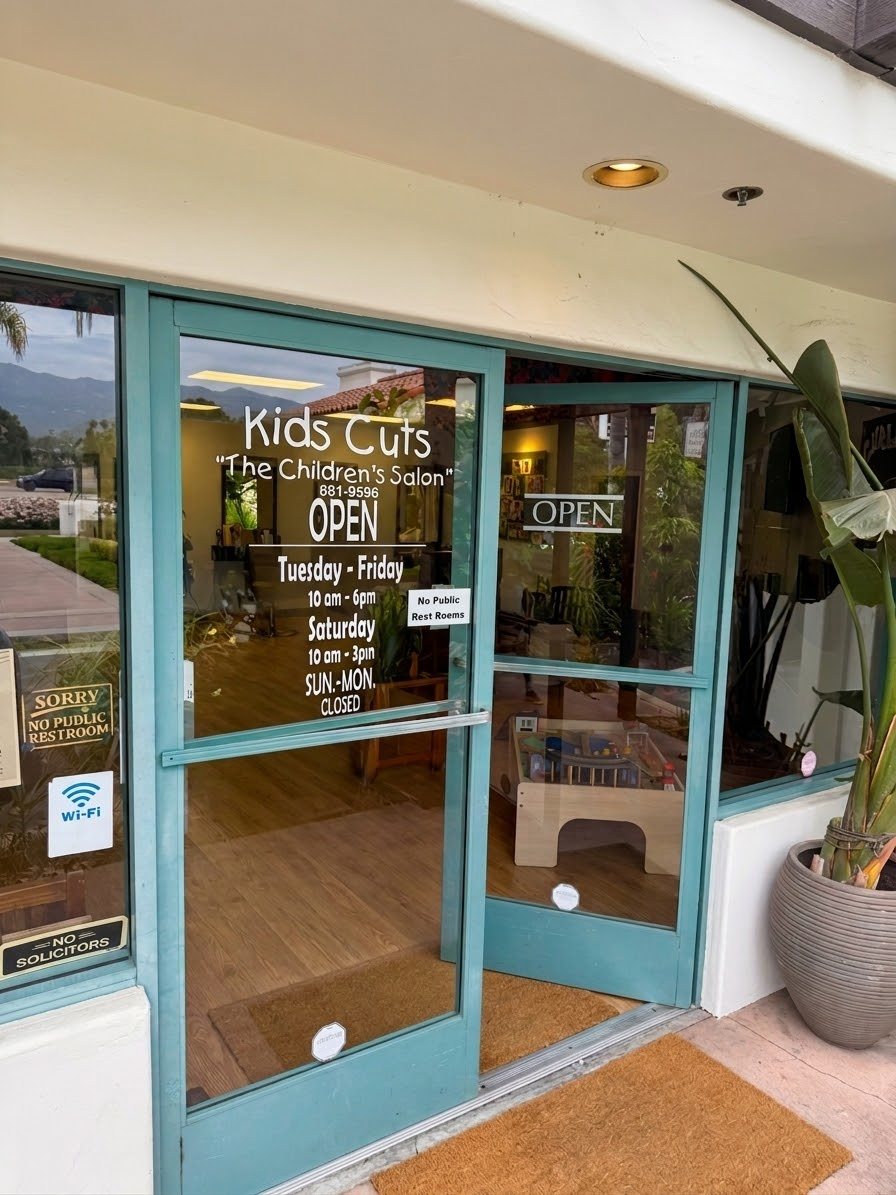 The teal front doors of Kids Cuts with the salon name, phone number and opening hours painted on the glass.