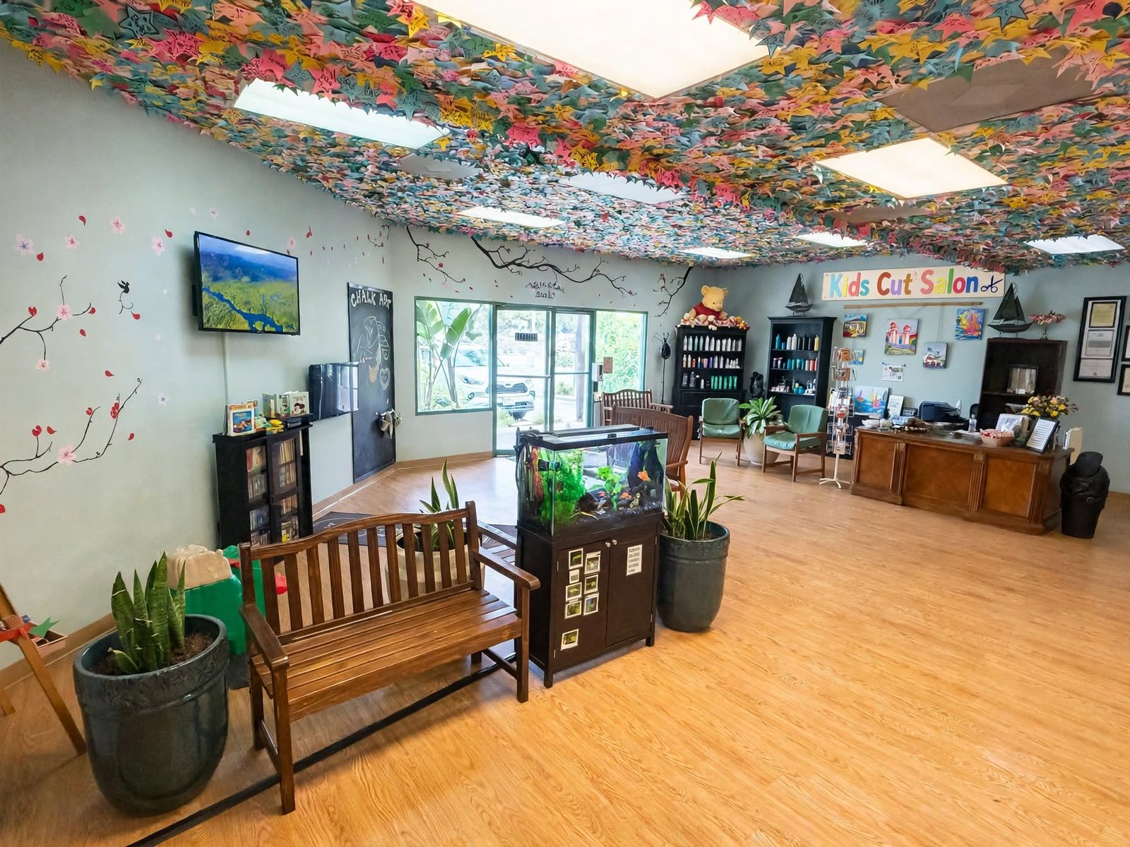The Kids Cuts waiting area with a wooden bench, aquarium, bookshelf, chalk art board, painted cherry blossoms on the wall and the hand-painted Kids Cut Salon sign.